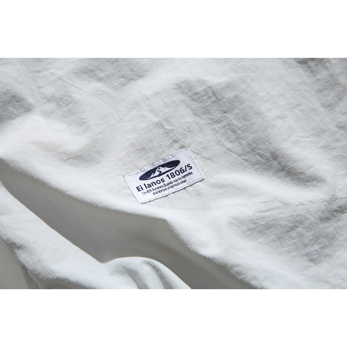 Close-up of a dark embroidered label on the pale gray shirt, showing the English text “El lanos 1806/S.”