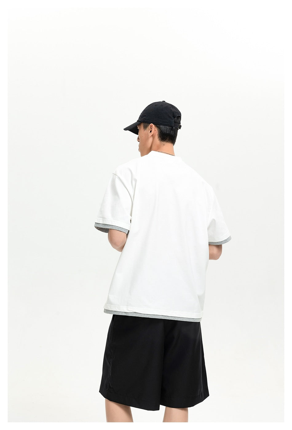 Rear view of a model wearing a white shirt with gray sleeve and hem trim, black shorts, and a black cap.