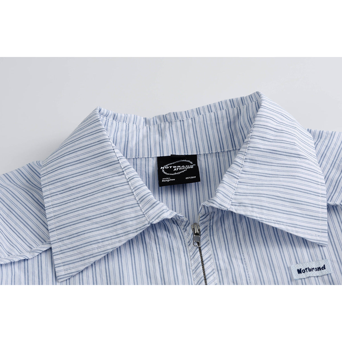 Close-up of a white shirt with fine blue stripes, a wide collar, front zipper, and sewn labels.
