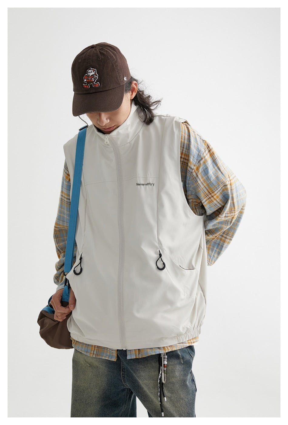 Front view of a model wearing an off-white sleeveless zip vest with drawcord pockets over a blue and ochre plaid shirt.