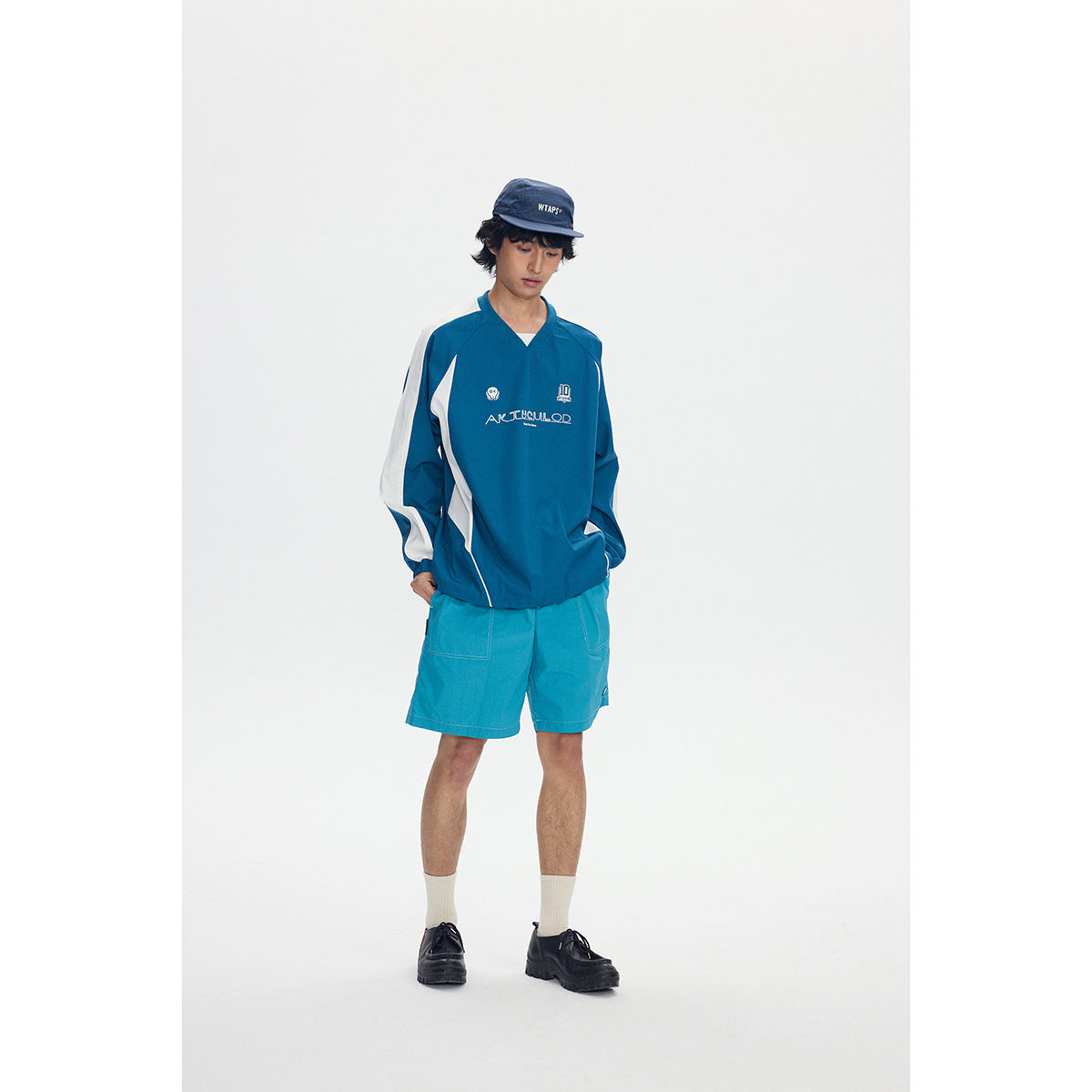Front view of a model wearing a blue-green V-neck pullover with white sleeve panels and teal shorts.