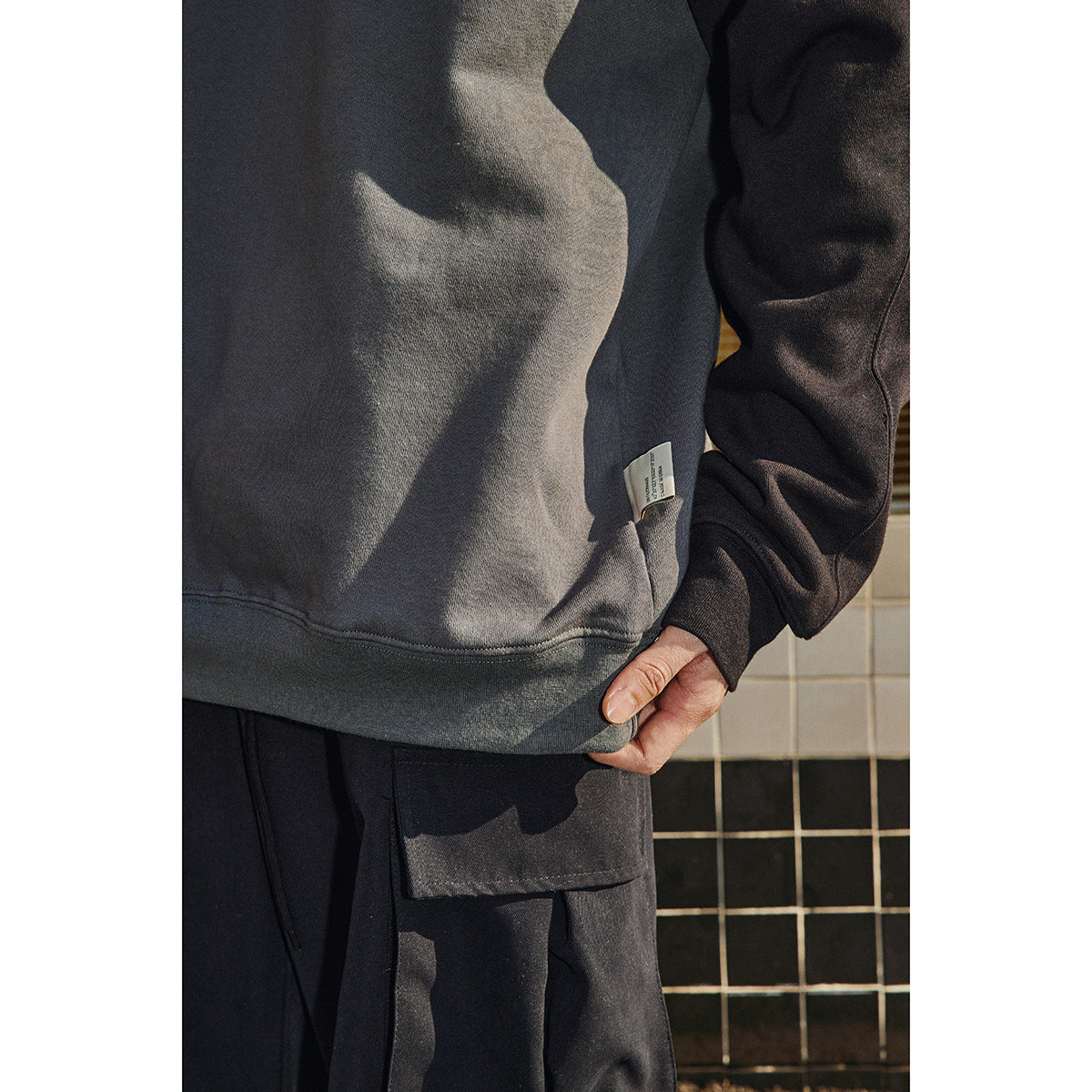 Close-up of a charcoal sweatshirt hem and side tag, with a hand holding it above black cargo pants.