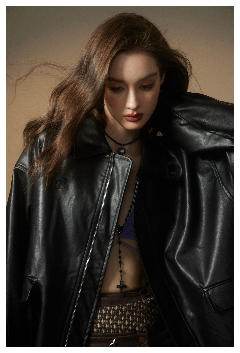 Close view of a model wearing a glossy black jacket, showing its pointed collar, front zipper, and small embroidered chest