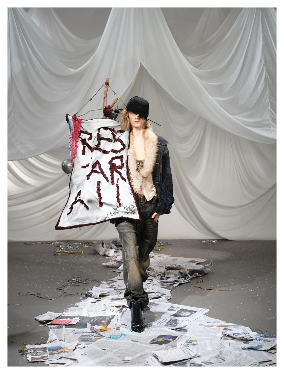Model in a charcoal fur-collar jacket, partly obscured by a white banner with fragmented dark red lettering.