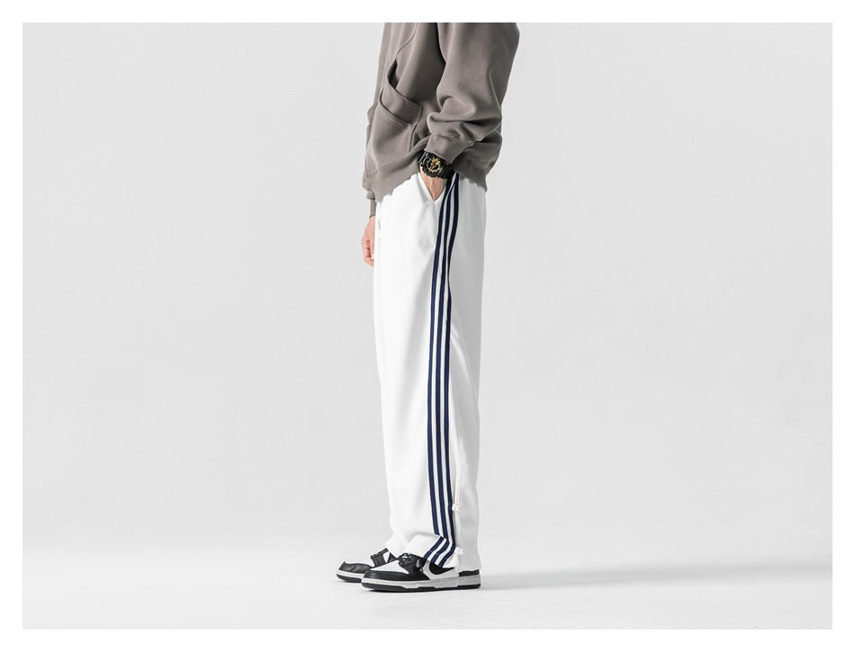 Side view of a model wearing loose white track pants with three navy side stripes and snap hems.