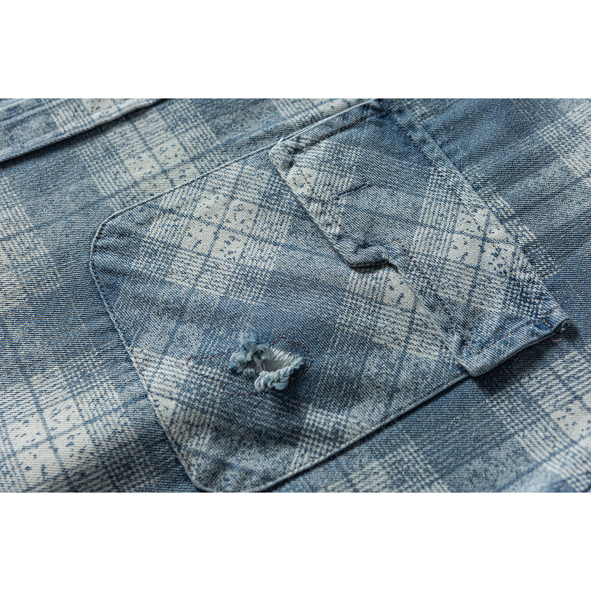 Close-up of a chest pocket on blue-gray patterned denim, showing a small distressed tear and buttoned flap.
