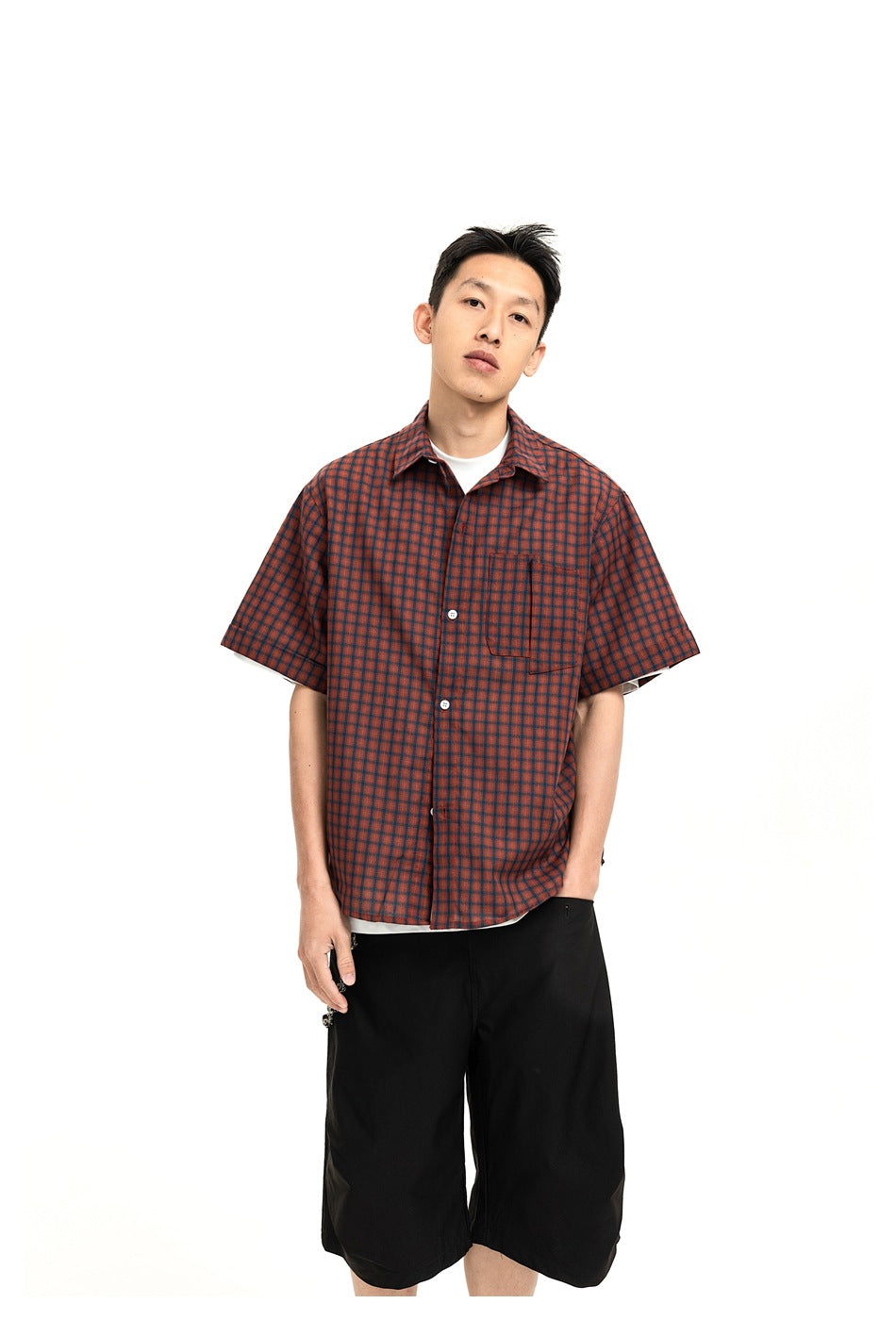 Model wearing a rust-red and dark check short-sleeve button-up shirt with a chest pocket and black shorts.