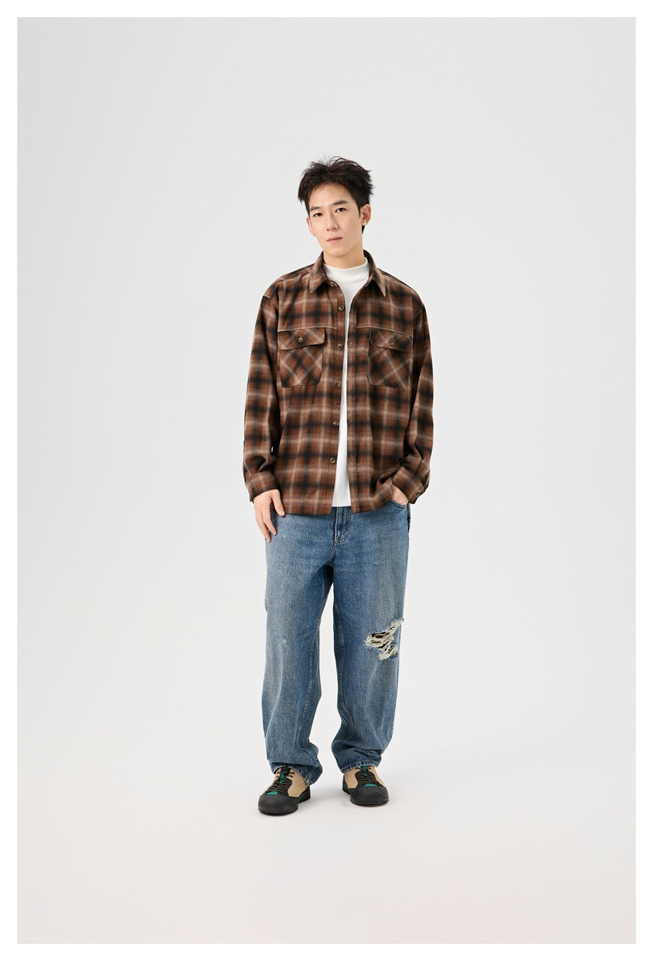 Man wearing a brown plaid button-up shirt open over a white top with blue jeans, full-length front view