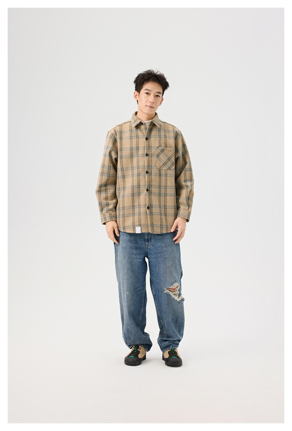 A man models a tan plaid button-up overshirt with a chest pocket and loose blue jeans.