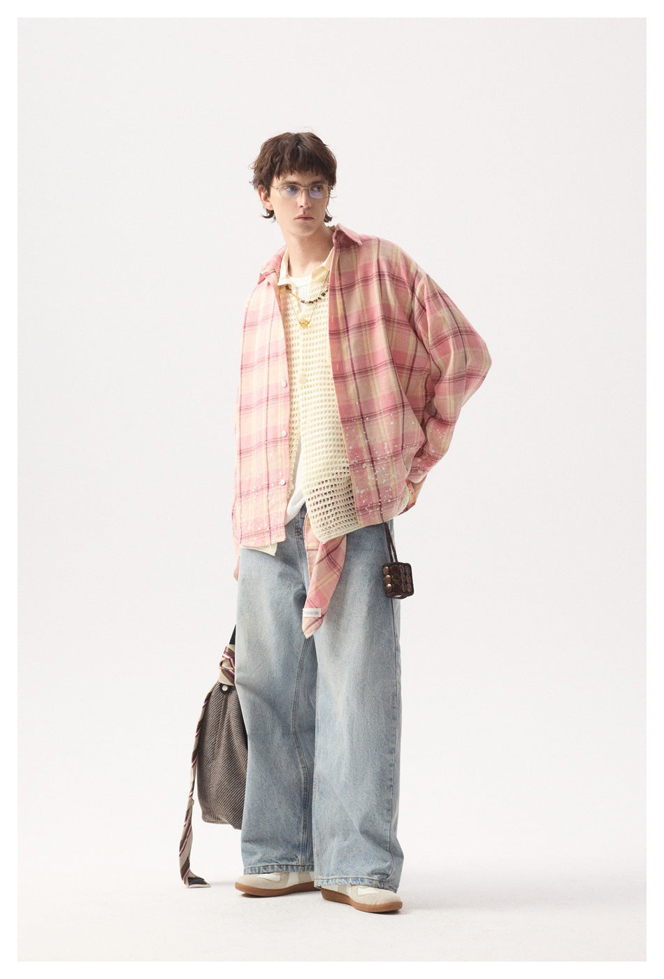 Full-length model in a pink plaid overshirt with white splatter details, cream knit vest, and loose light-wash jeans.