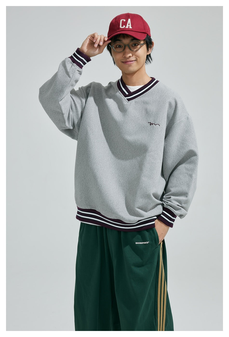 Smiling model adjusts a red cap while wearing a gray striped-trim V-neck sweatshirt and dark green pants.