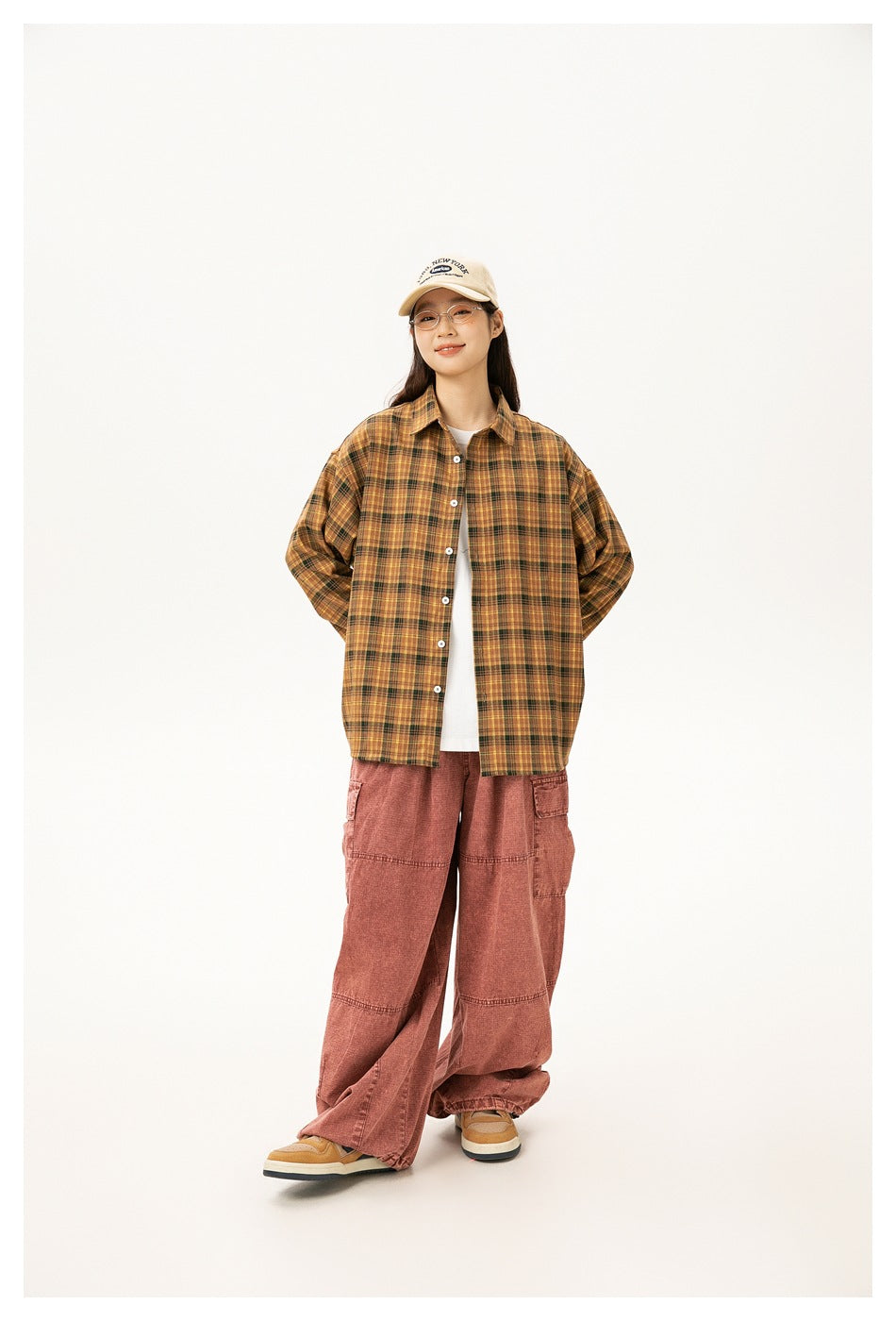 Full-length view of a model in a mustard plaid button-up shirt, rust cargo pants, sneakers, and a beige cap.