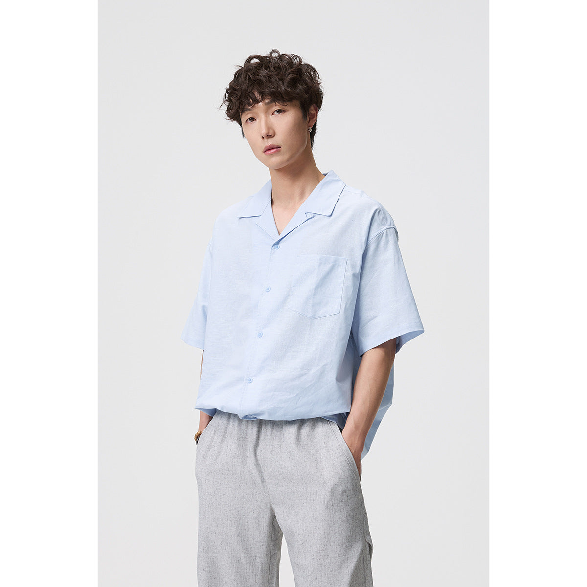 Model wearing a light blue short-sleeve button-up shirt with a notched collar and chest pocket, paired with gray trousers.