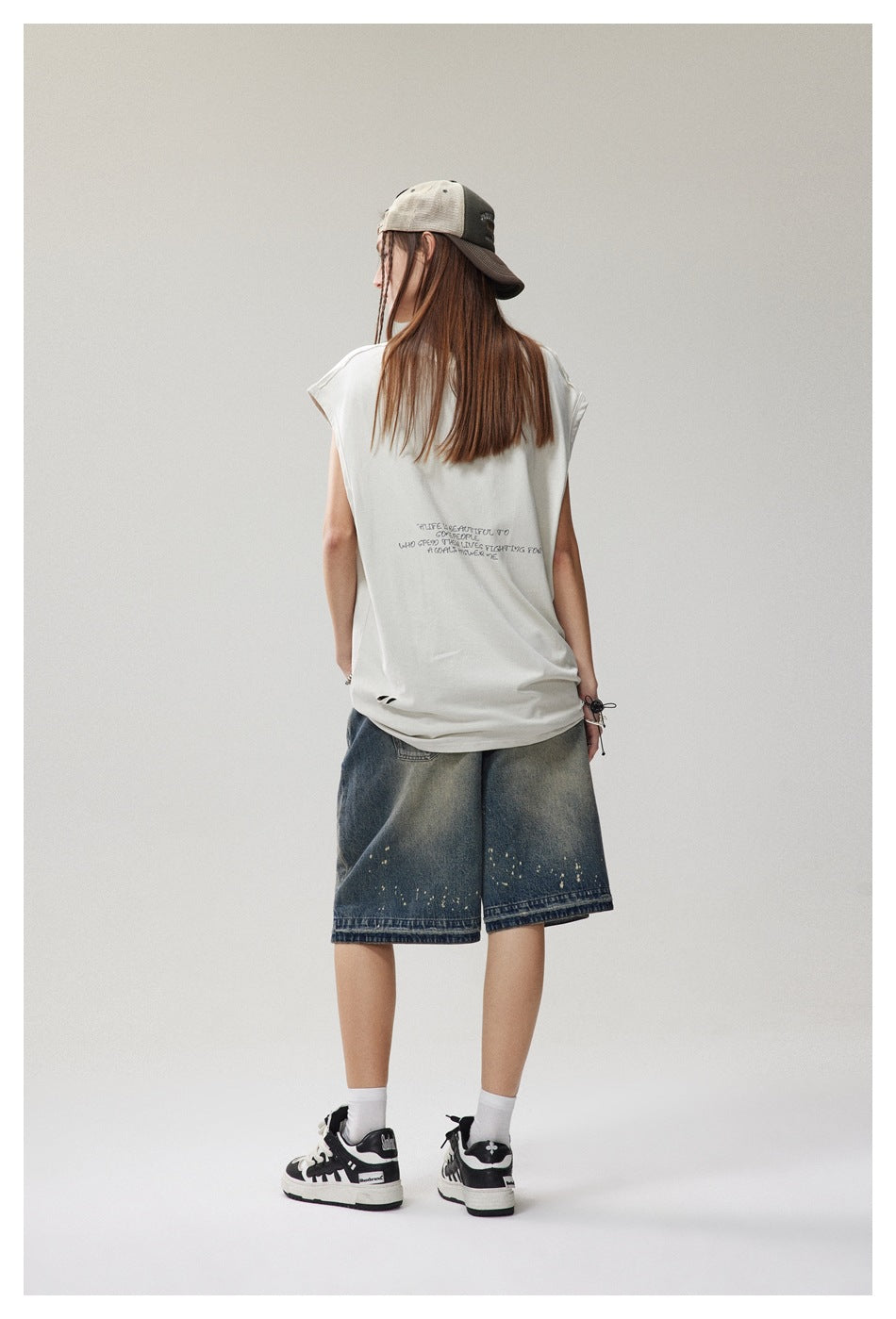 Back view of model in a light beige sleeveless top with small printed text, denim shorts and sneakers