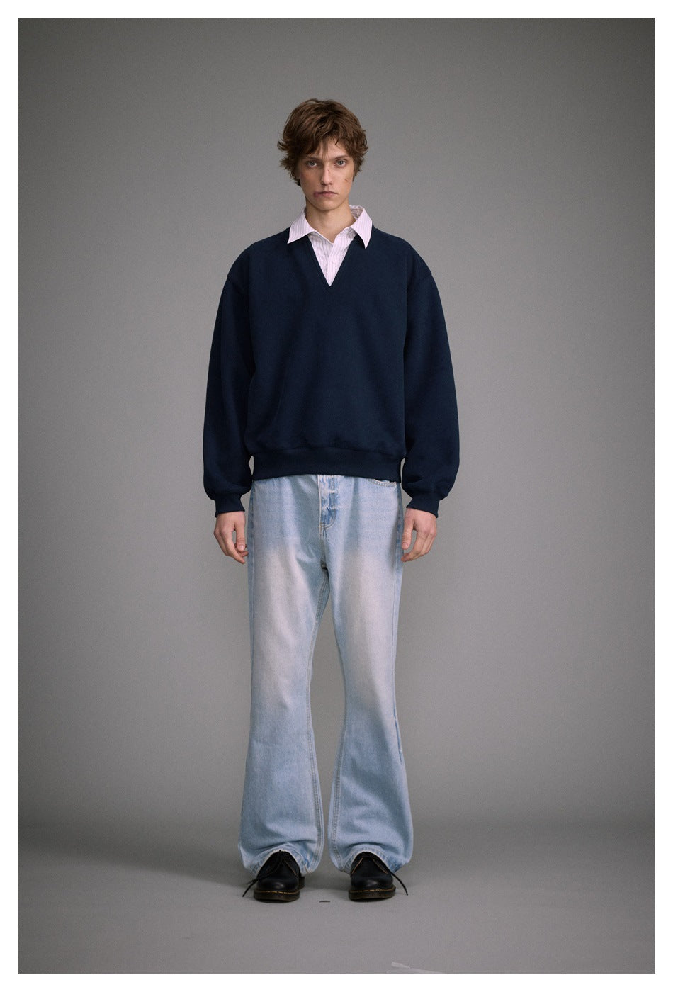 Full-length front view of a model in a navy V-neck sweatshirt, pale pink striped shirt, and light-wash wide-leg jeans.