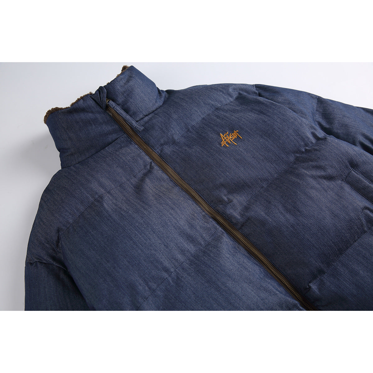 Close-up of a navy padded jacket front with a brown zipper, high collar, and small gold embroidered chest mark.