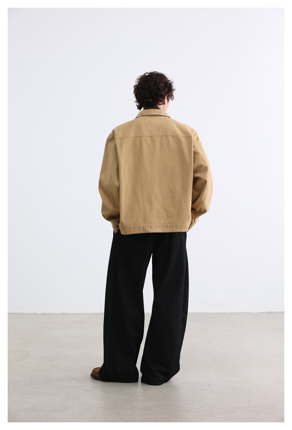 Rear view of a model wearing a tan jacket with multicolor collar trim and black wide-leg trousers.
