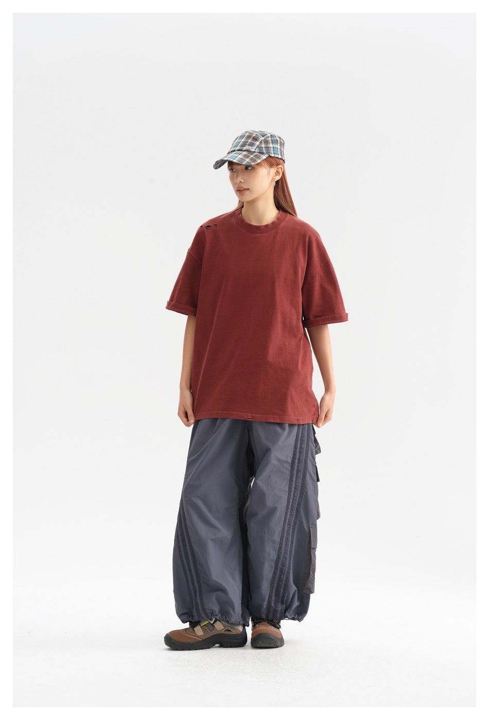Model wearing an oversized brick-red T-shirt with small shoulder distressing, gray cargo pants, and a plaid cap.