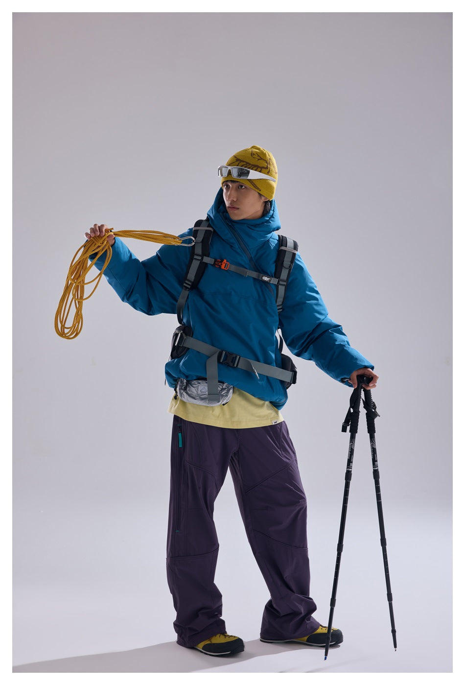 Model wearing a bright blue hooded pullover and purple trousers with a climbing harness, rope and trekking poles.