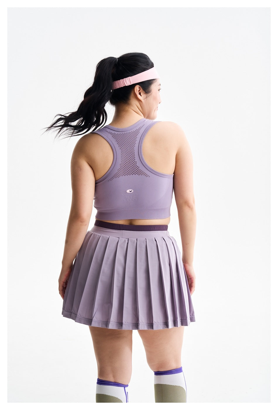 Rear view of a model in a lavender racerback sports bra and pleated skirt, showing the perforated back panel.