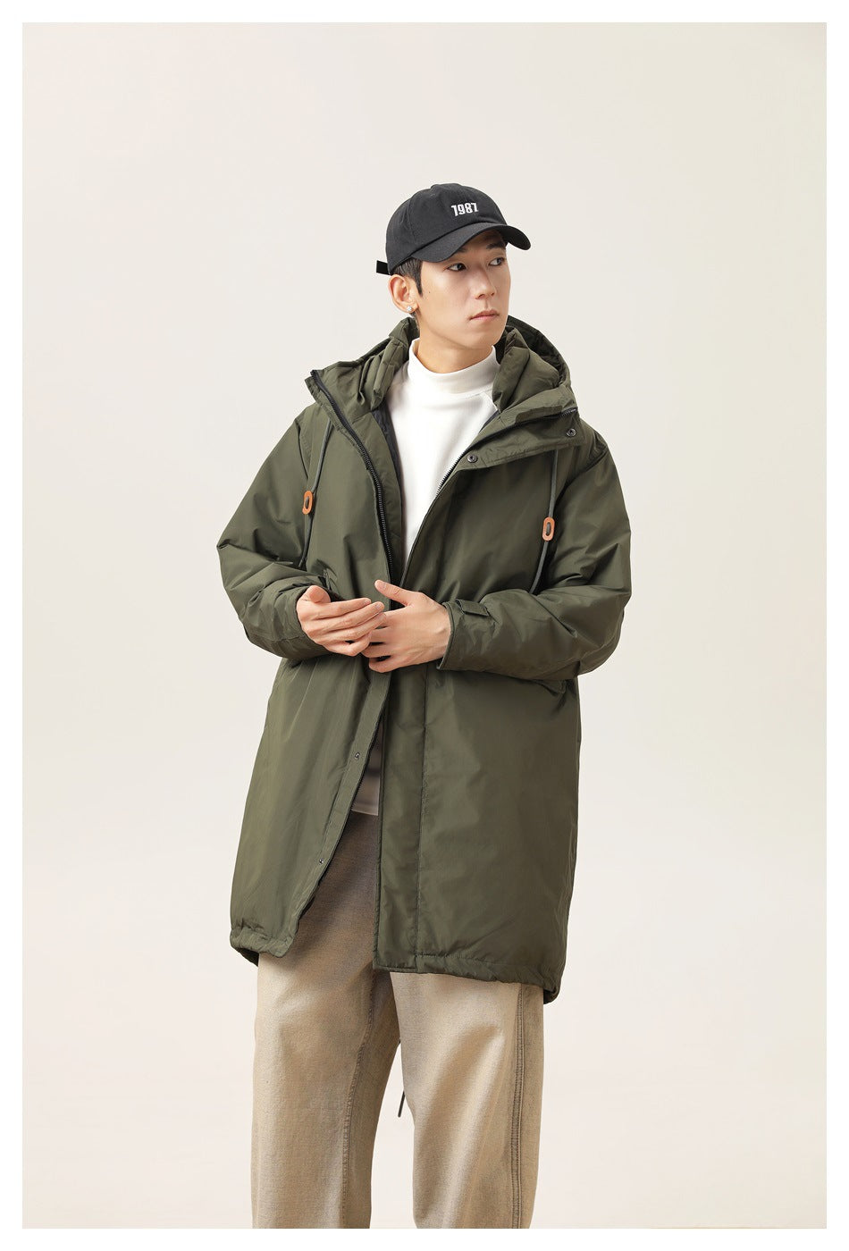 Model wearing an army green long hooded jacket partly unzipped over a white mock-neck top and tan pants