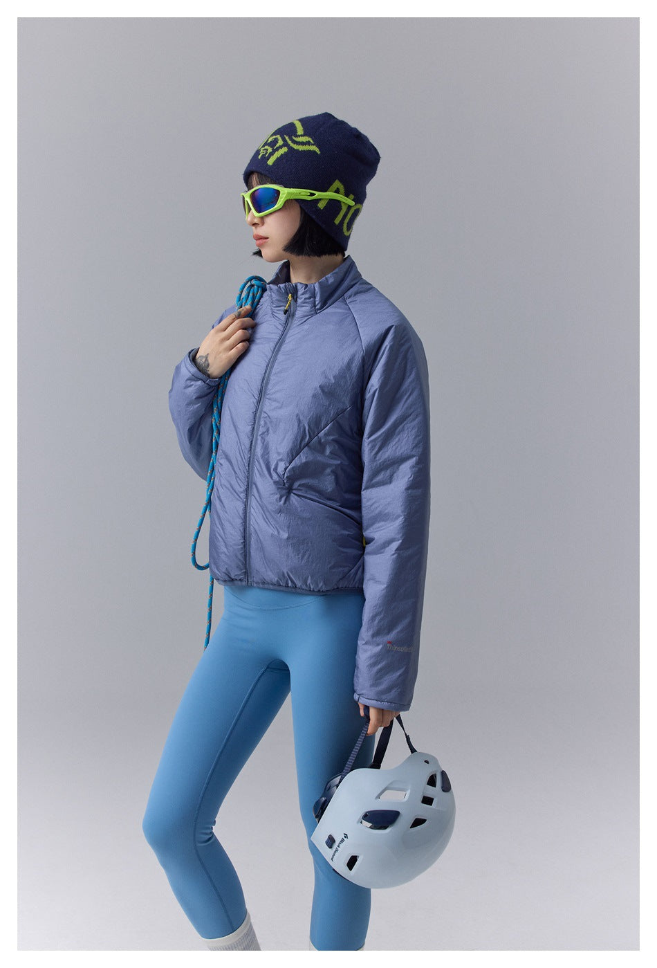 Model in a periwinkle-blue padded zip jacket and blue leggings, holding a climbing helmet and rope.