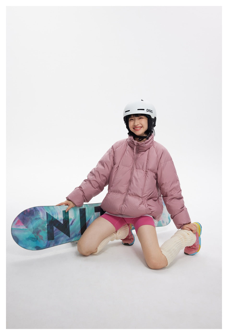 Model seated on a colorful snowboard, wearing a dusty pink puffer jacket and pink shorts.