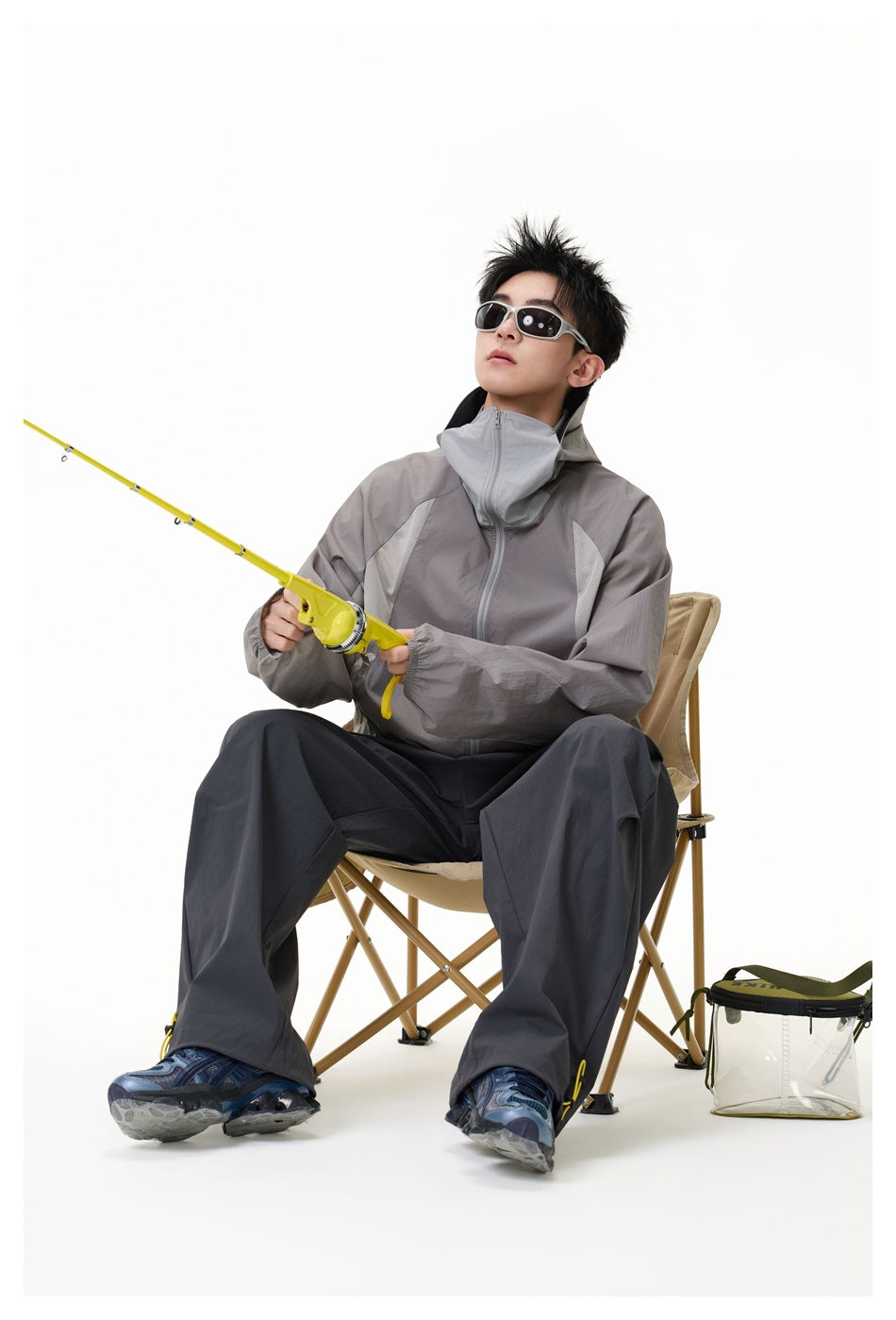 Seated model wearing the gray hooded zip jacket with raised collar, dark wide pants and sunglasses, holding a yellow fishing