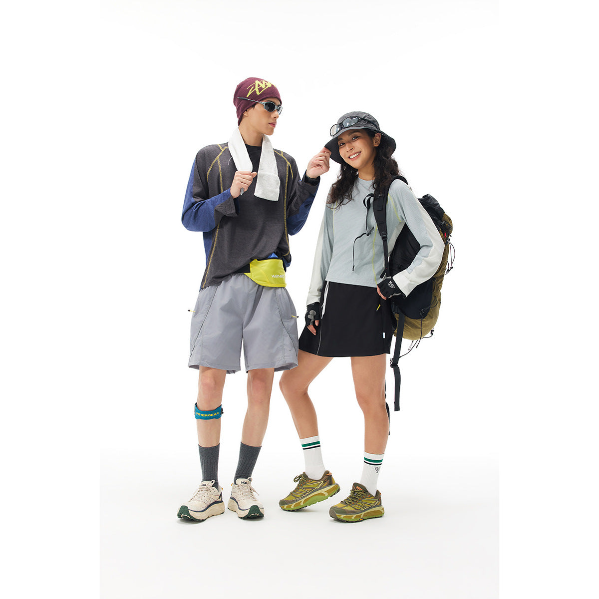 Two hikers pose in color-block long-sleeve tops, shorts and skirts, with hats, backpacks and trail shoes.