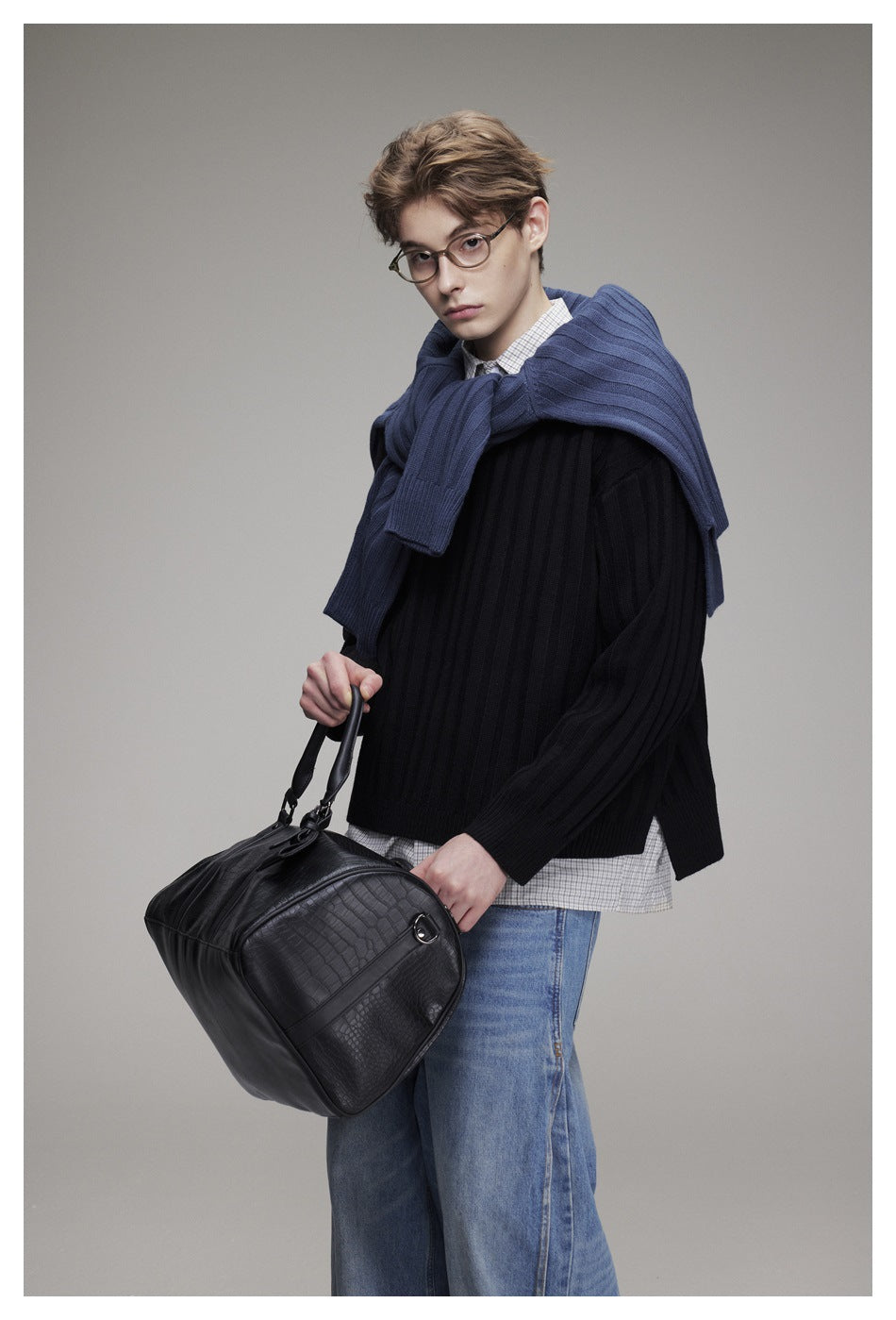 Closer view of a model wearing a black ribbed sweater over a checked shirt, blue sweater draped over his shoulders, and