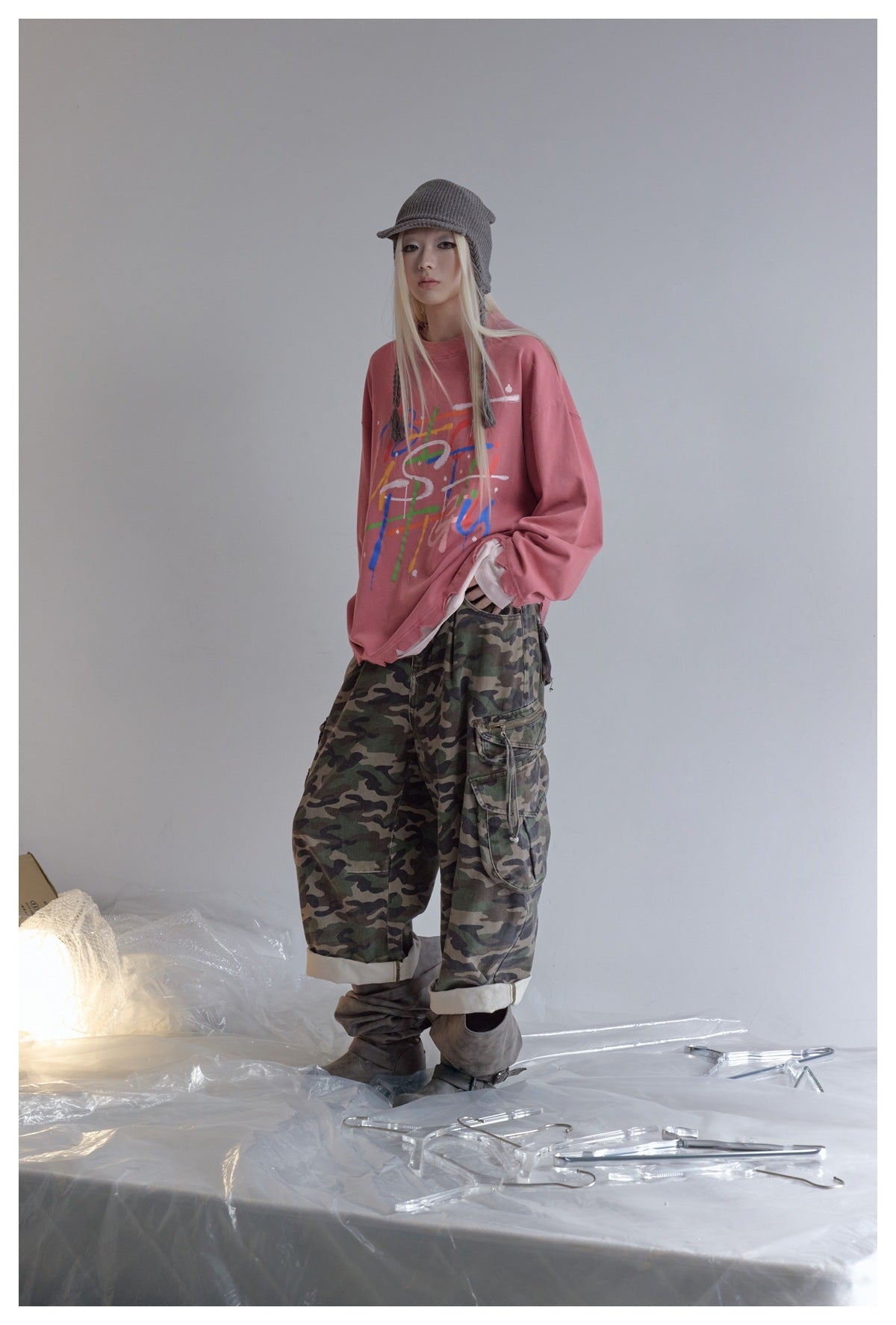 Model wearing oversized pink graffiti-print long-sleeve top with camouflage cargo pants, knit cap and gray boots