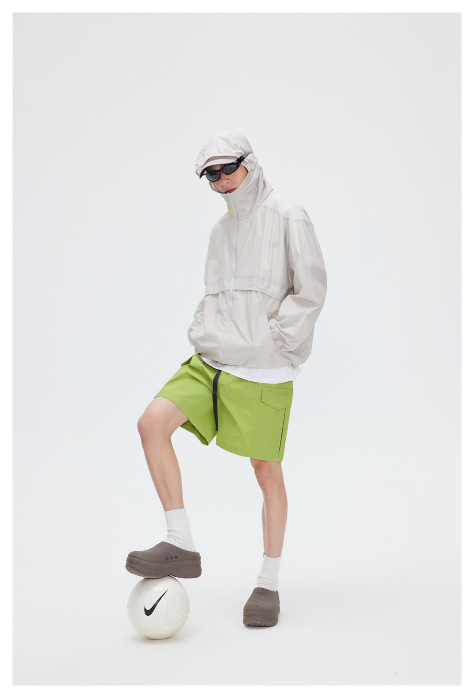Full-length model wearing a pale beige hooded jacket, lime-green shorts, a cap and sunglasses.