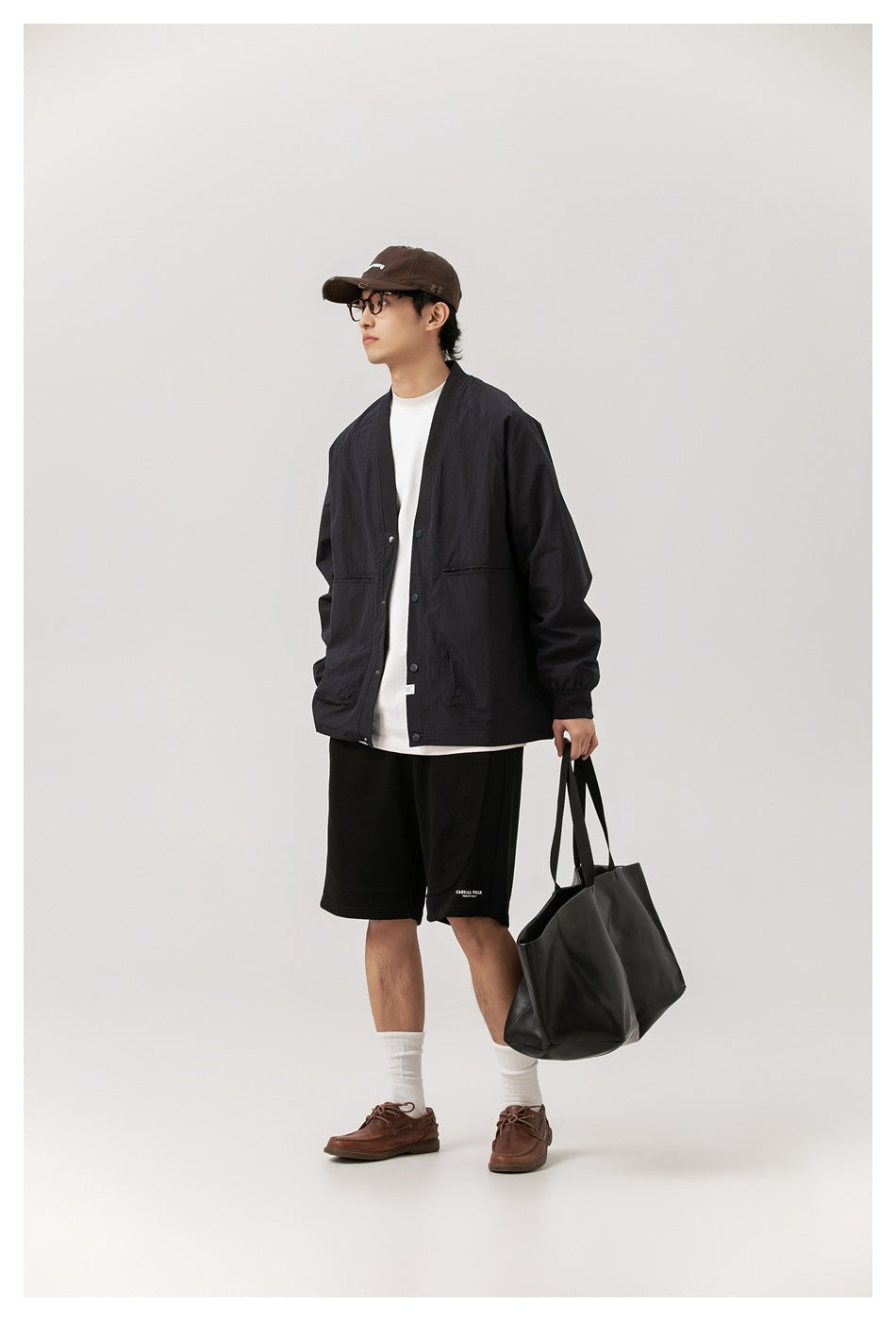 Full-length model wearing a black snap-front jacket over a white shirt, with black shorts and a brown cap.