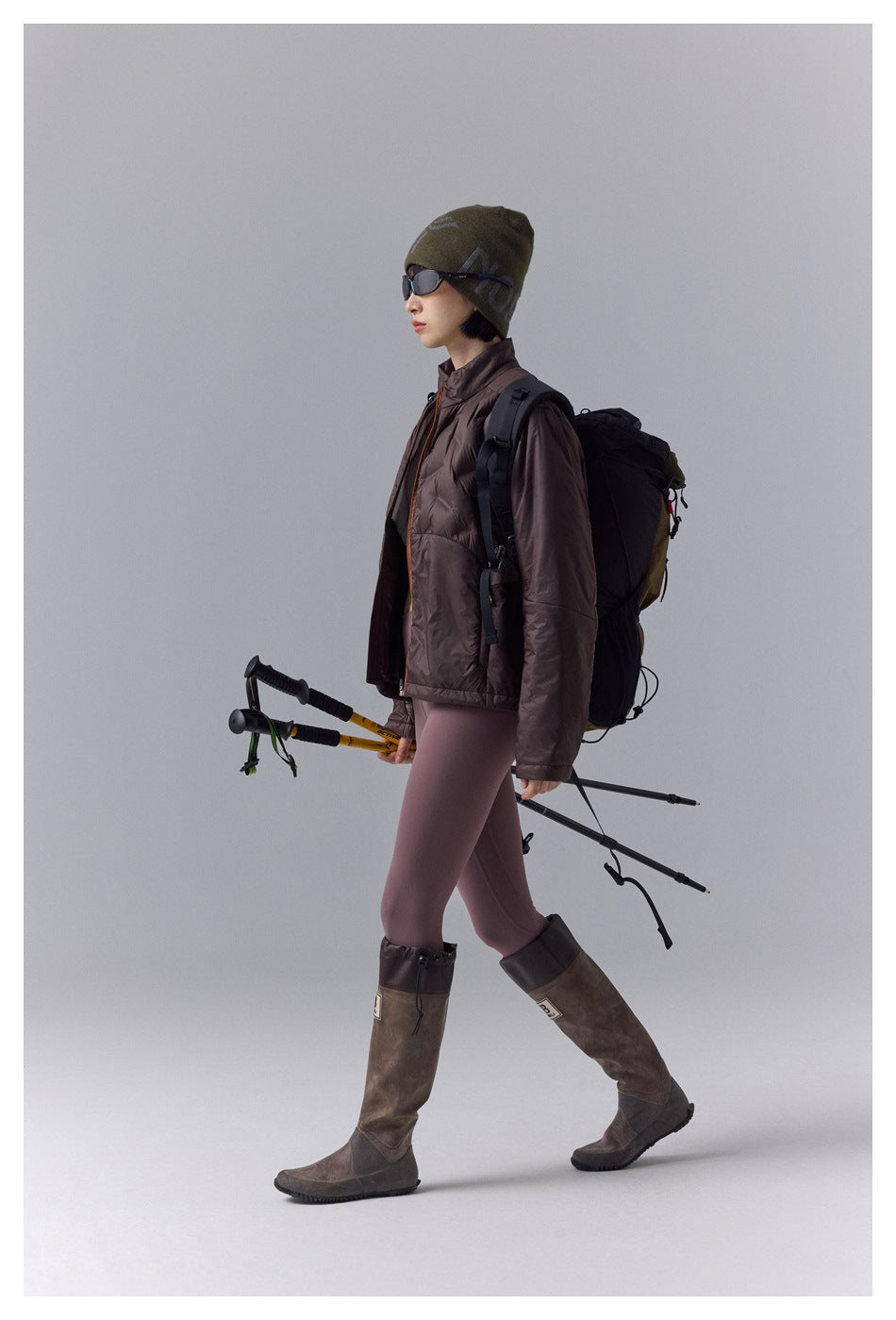 Side view of a model wearing a brown puffer jacket, mauve leggings, tall boots, and an olive beanie.