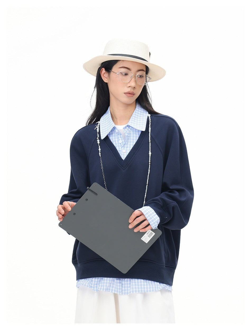 Close-up of model in navy V-neck sweatshirt with blue checked collar, holding a gray clipboard, wearing hat and glasses