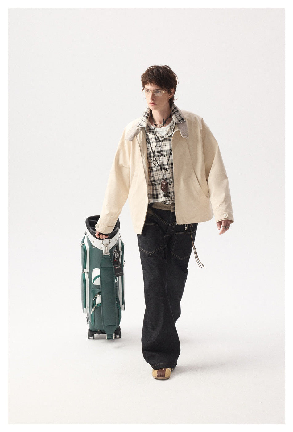 Model wearing a beige zip jacket over a plaid shirt and dark jeans, walking with a green golf bag
