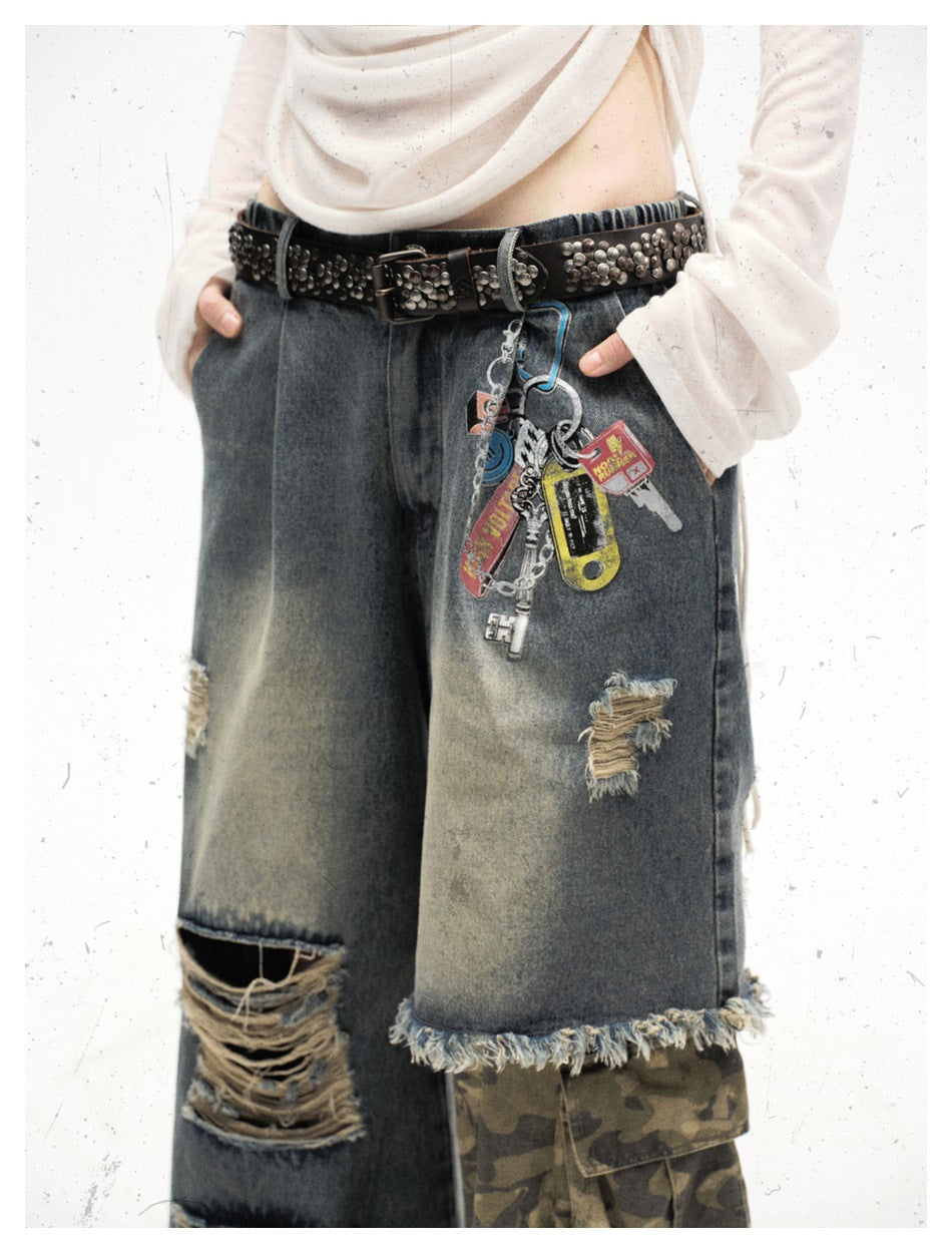 Front view of a model wearing distressed wide-leg denim pants with a camouflage cargo panel, frayed hems and dangling charms.