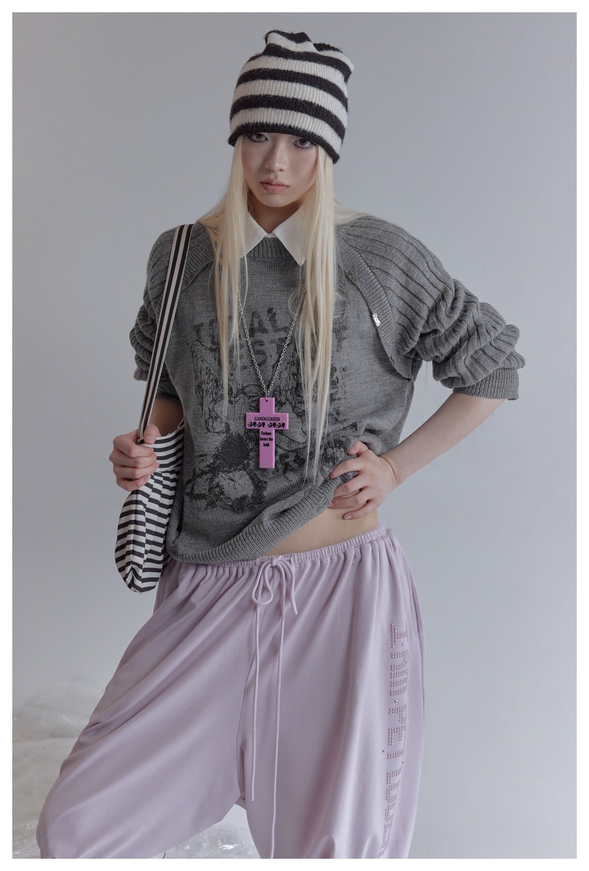 Model wearing a loose gray graphic raglan sweater with lilac drawstring pants, a striped beanie, and a striped shoulder bag.