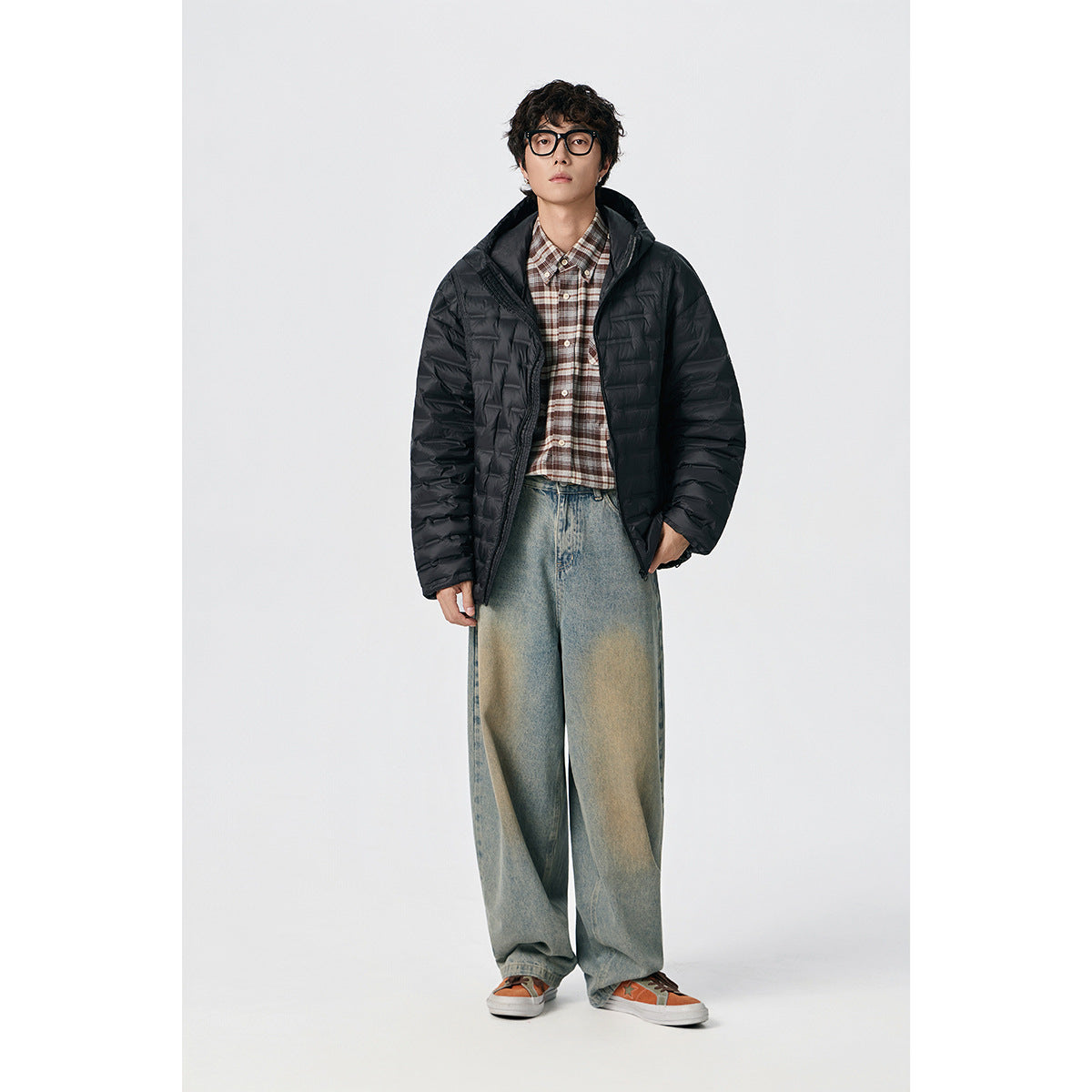 Model wearing a black hooded quilted jacket over a plaid shirt, shown full length against a light background.