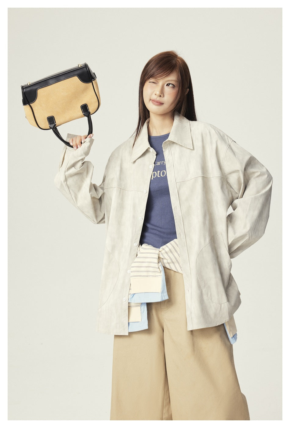 Model in a light beige oversized shirt jacket, blue top, and tan wide-leg trousers, holding a two-tone bag.
