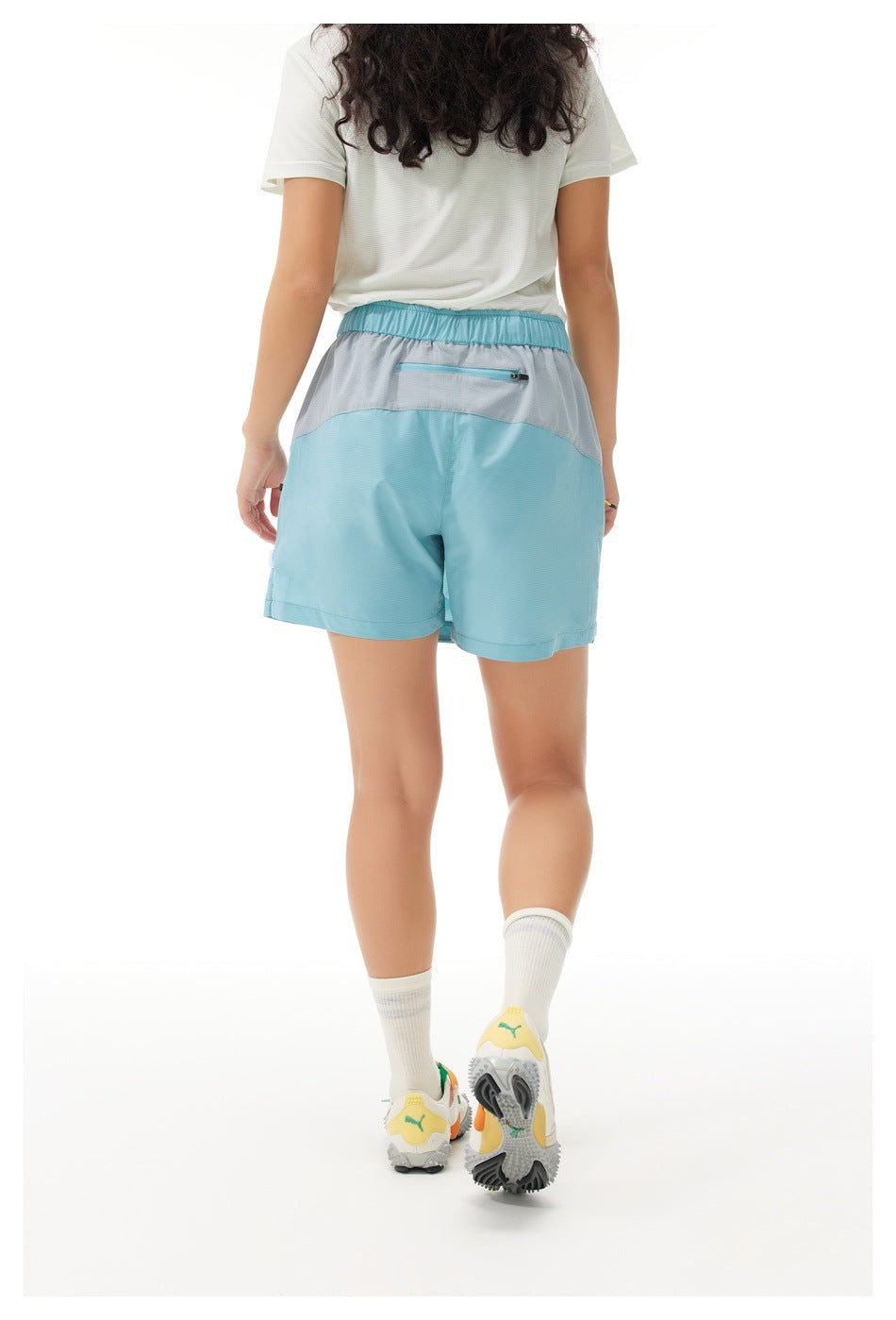 Rear view of a model wearing blue athletic shorts with a gray curved waistband panel and zip pocket.