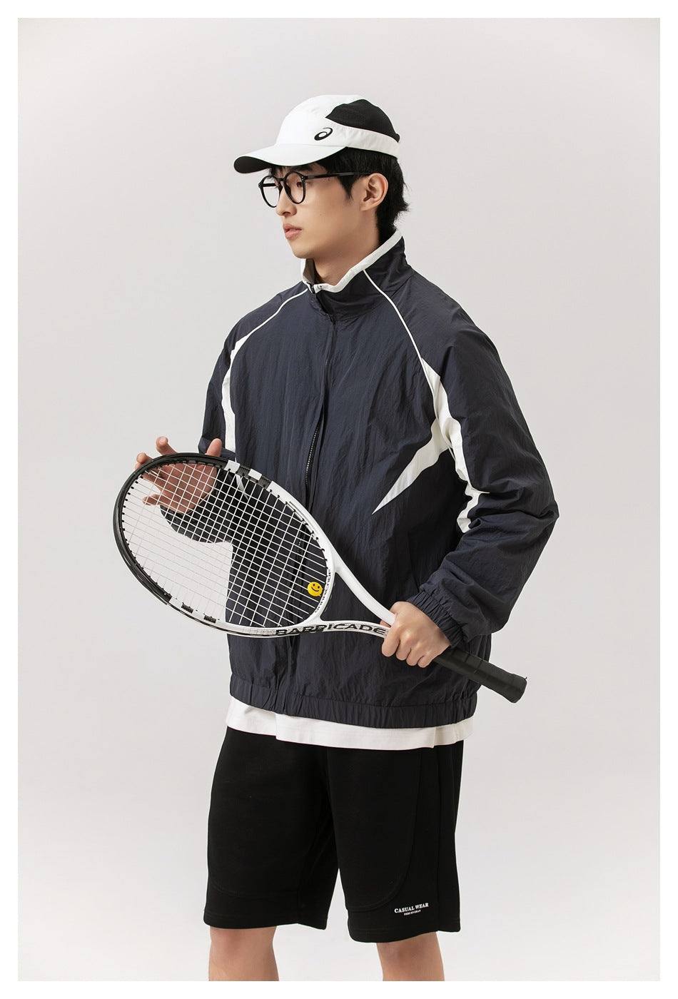Model wearing a dark navy zip jacket with white panels and black shorts, holding a tennis racket across the front.