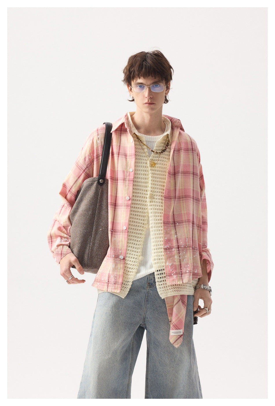 Front view of a model wearing a pink plaid overshirt with white splatter details over a cream knit vest and light-wash jeans.