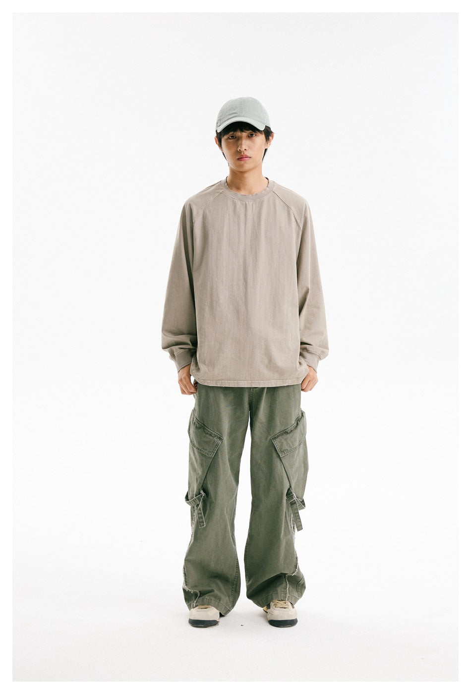 Full-length front view of a model wearing a loose taupe crewneck, olive cargo pants, and a light cap.
