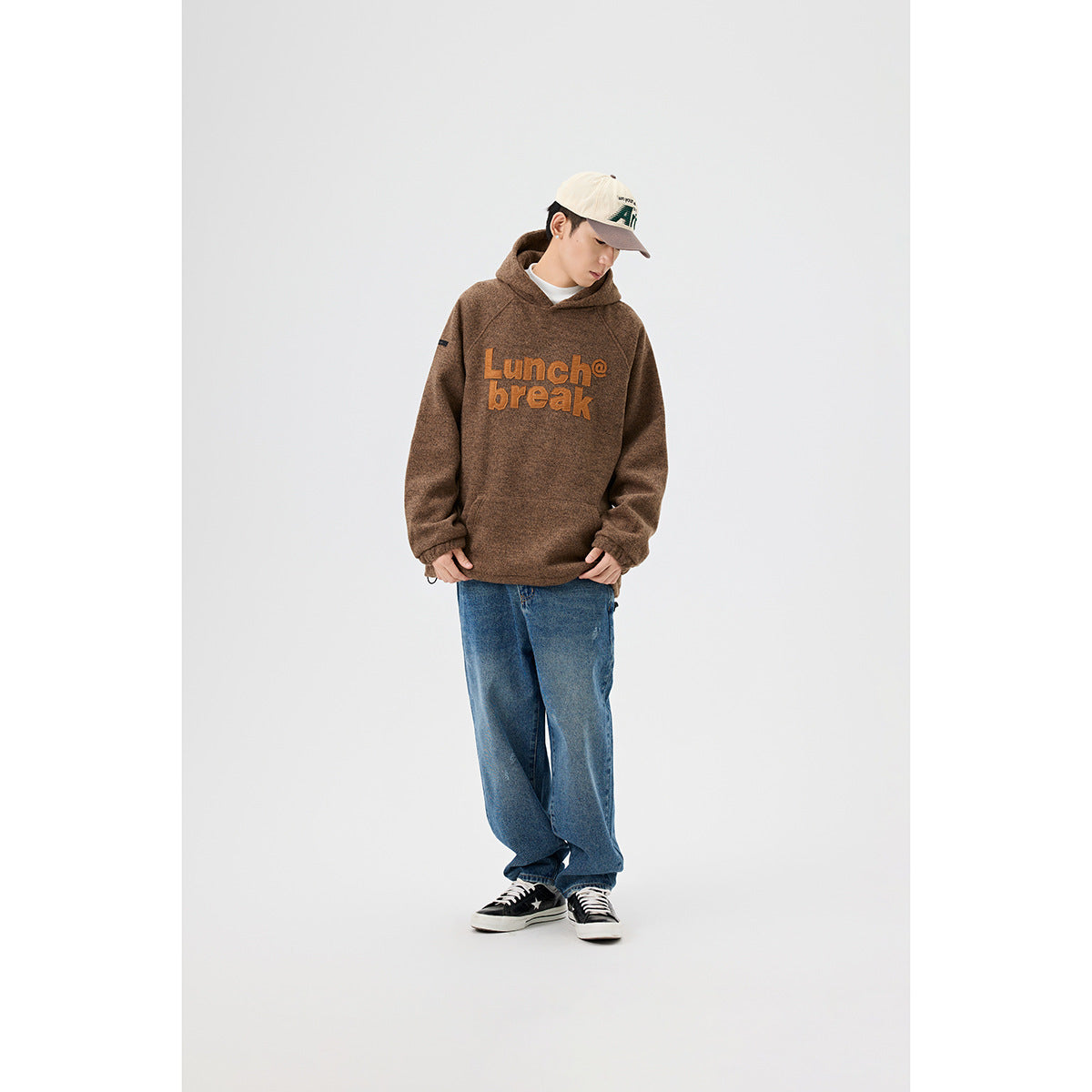 Model wearing a brown hooded pullover with orange “Lunch break” lettering, blue jeans, and a cream cap.