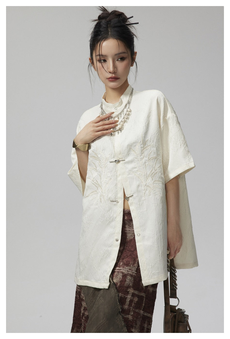 Model wearing an ivory embroidered short-sleeve tunic with a stand collar and silver frog closures.