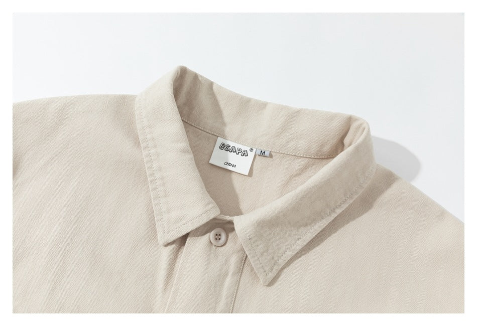 Close-up of a cream herringbone shirt collar, top button, and sewn neck label, framed by a white border.