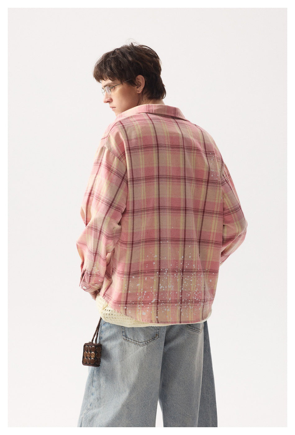 Rear view of a model wearing a pink plaid overshirt with white splatter details and loose light-wash jeans.