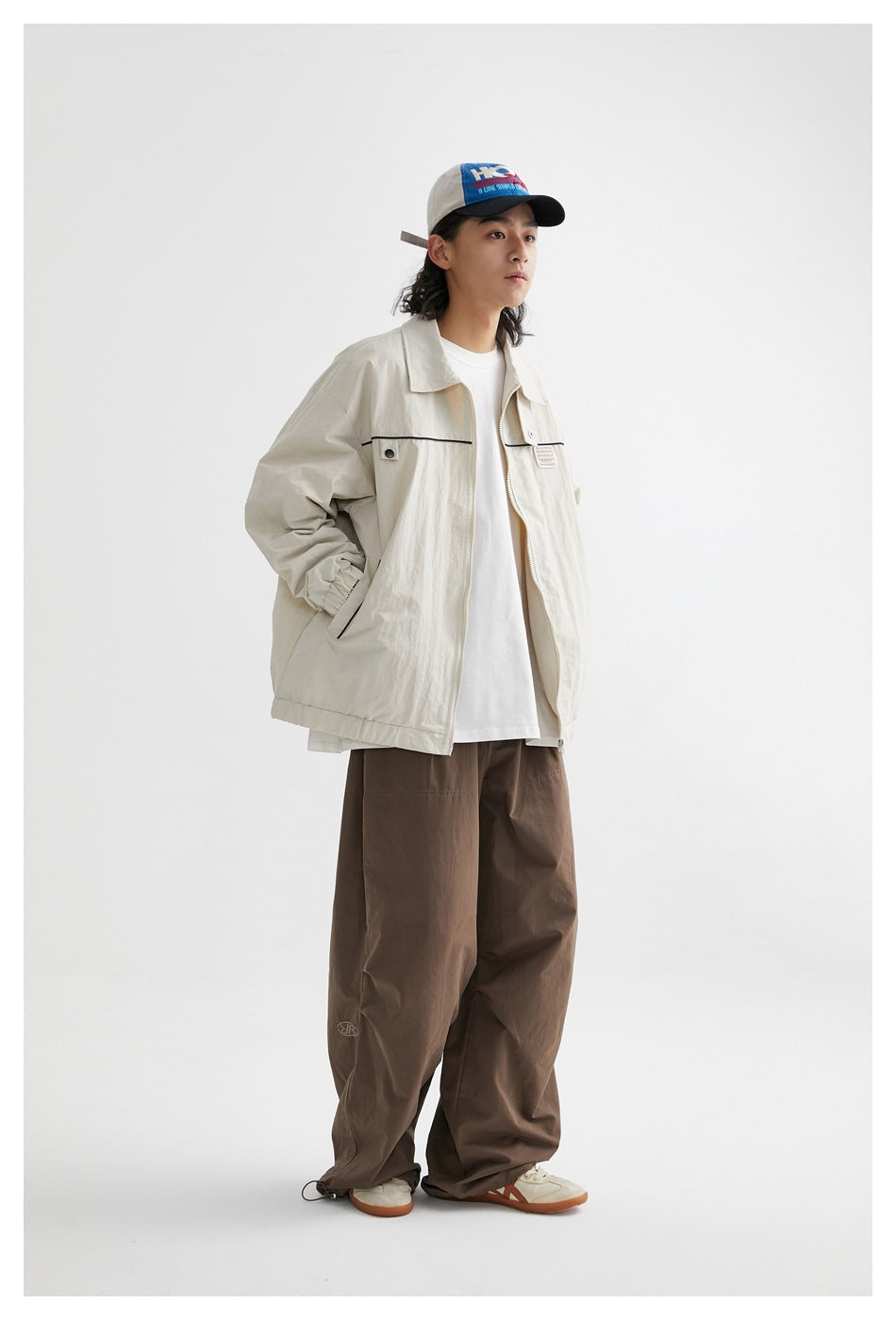 Model wearing an open beige zip jacket over a white tee with brown wide-leg pants and a blue and beige cap, full length