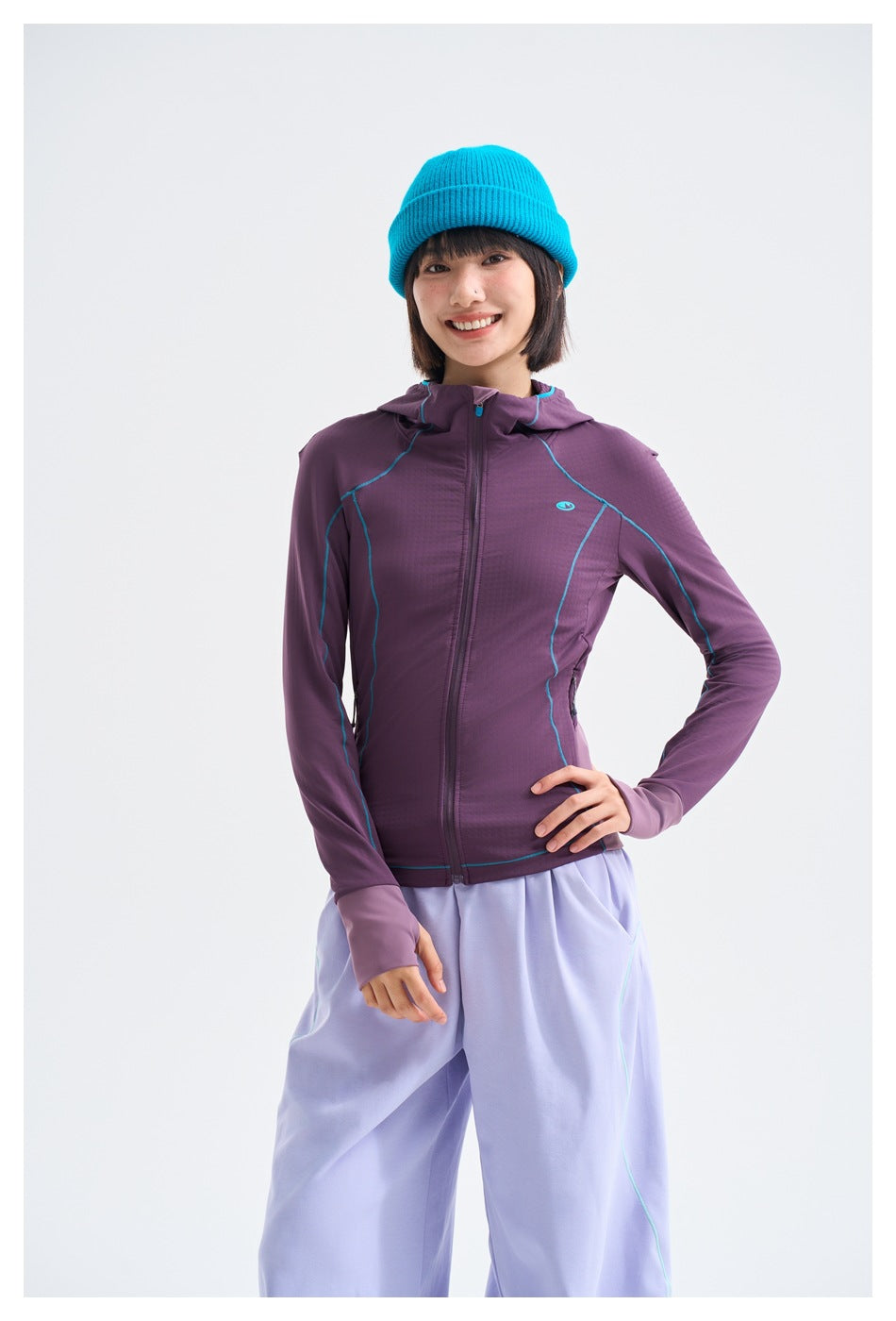 Model wearing a purple hooded zip jacket with turquoise seam piping and light lavender pants, plus a bright turquoise beanie.